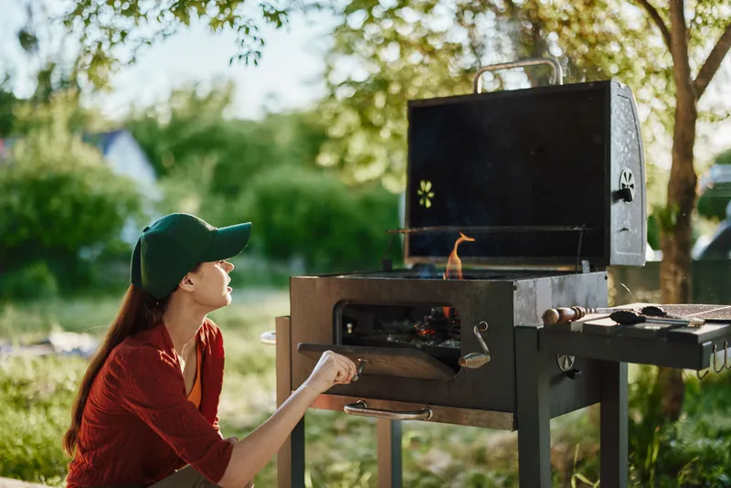 Young woman preparing barbecue on a sunny day, wearing a cap and casual clothes, surrounded by greenery, highlighting outdoor cooking and summer vibes