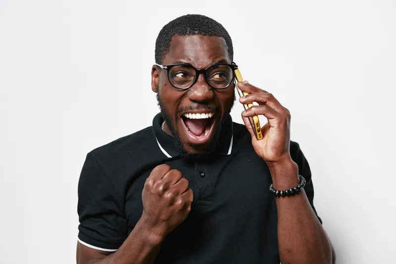 African American man with dark skin and glasses expressing excitement while talking on smartphone, wearing black polo shirt and bracelet on white background.