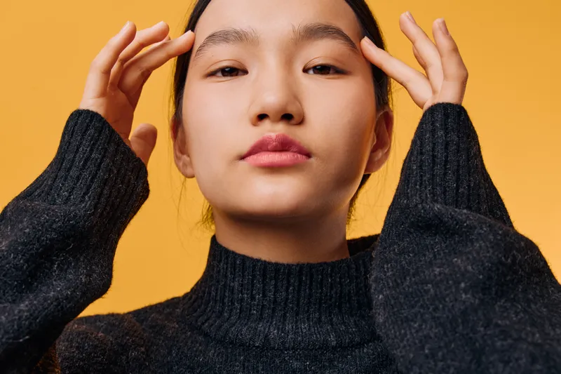 Young woman posing with hands near face wearing black sweater on orange background. Portrait of female with natural skin, minimal makeup, and serious expression for fashion or beauty.