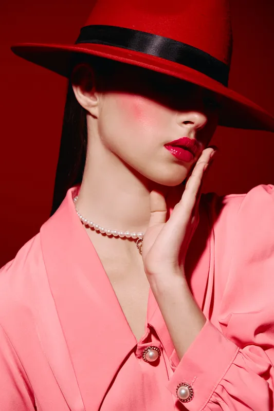 Woman in bright pink design outfit and red hat with glossy lips and pearl necklace poses with elegant hand near face. Fashion beauty concept with moody studio lighting.