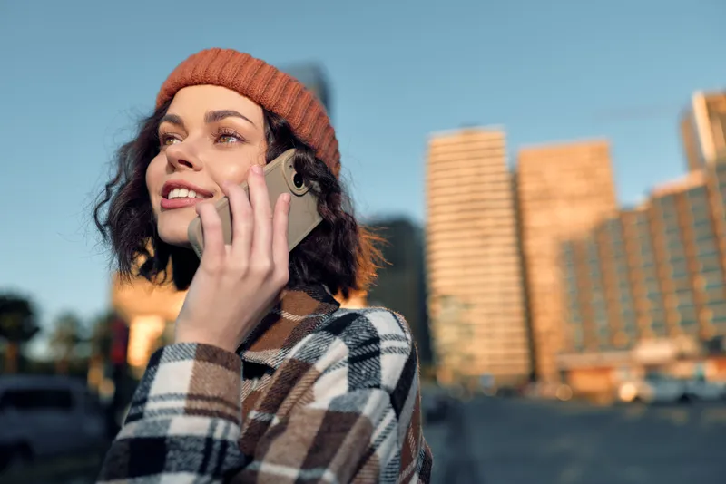 Woman on phone in city at golden hour, smiling in beanie and checkered coat; candid lifestyle portrait capturing authenticity, emotional storytelling and mindful living in urban glow.