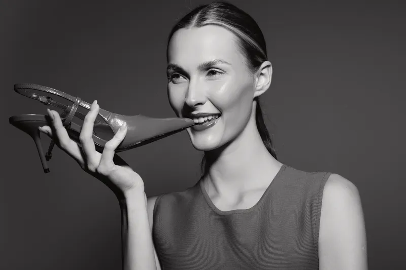 Smiling white woman with fair skin holding a high heel shoe near her mouth in studio. Concept for fashion, humor, and beauty with elegant style.