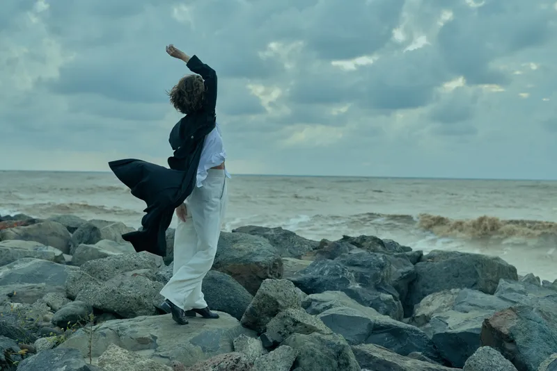 Couple dancing on rocky shoreline, windswept embrace, sea backdrop, flowing garment and white pants, dynamic movement against stormy ocean horizon