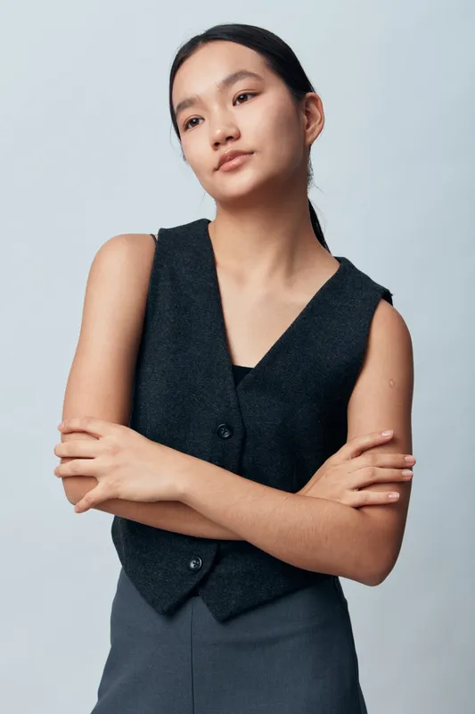 Young woman with long dark hair wearing sleeveless vest and pants posing with crossed arms on light background. Casual fashion, confident posture and thoughtful expression portrait in studio.