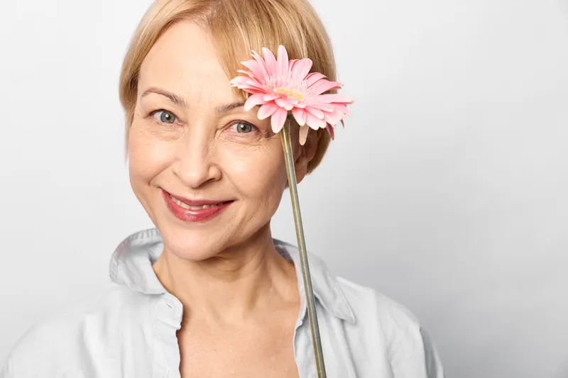Smiling elderly woman with pink flower near her face, studio portrait on white background. Beauty care concept with skincare and wellness, gentle grooming, soft light, calm today.
