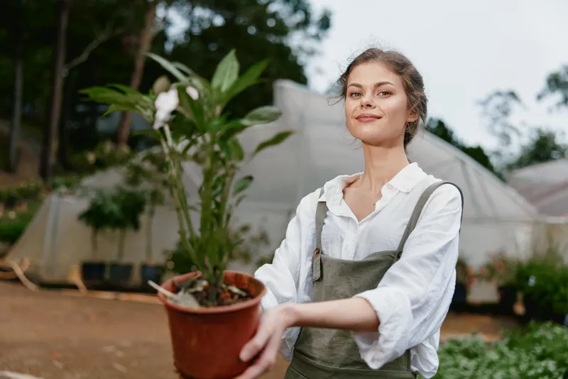 Young woman in a garden holding a potted plant, dressed in casual gardening attire, showcasing a joyful expression surrounded by greenery