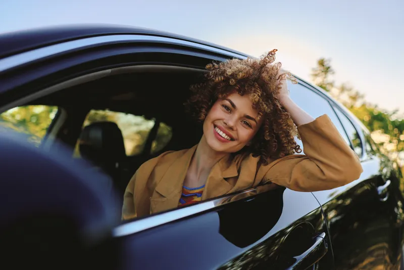 A smiling woman sits in a glossy car with sunlit retro styling, leaning against the window for a casual, confident lifestyle moment in bright daylight. Natural expression. Warm tone.
