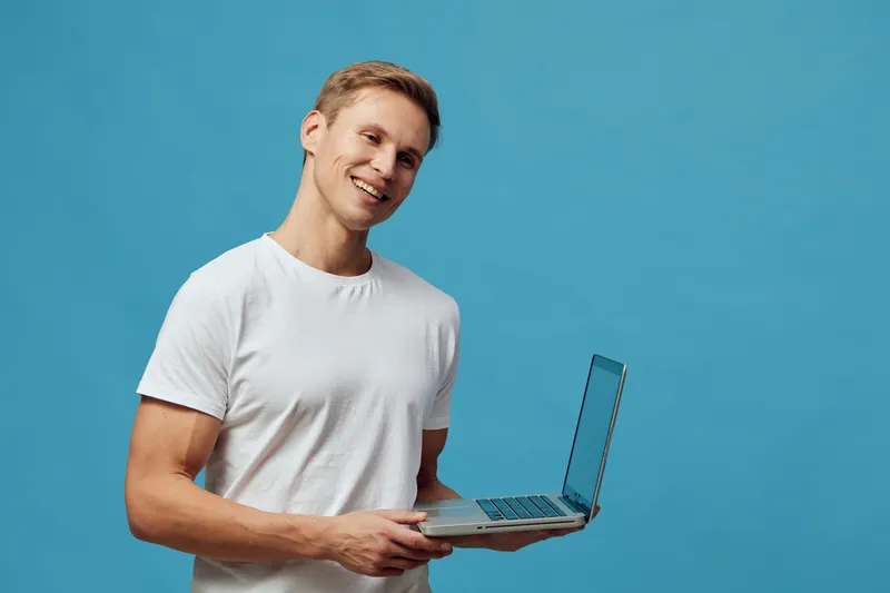 smiling young caucasian man wearing white t-shirt holding laptop working remote digital nomad blue background