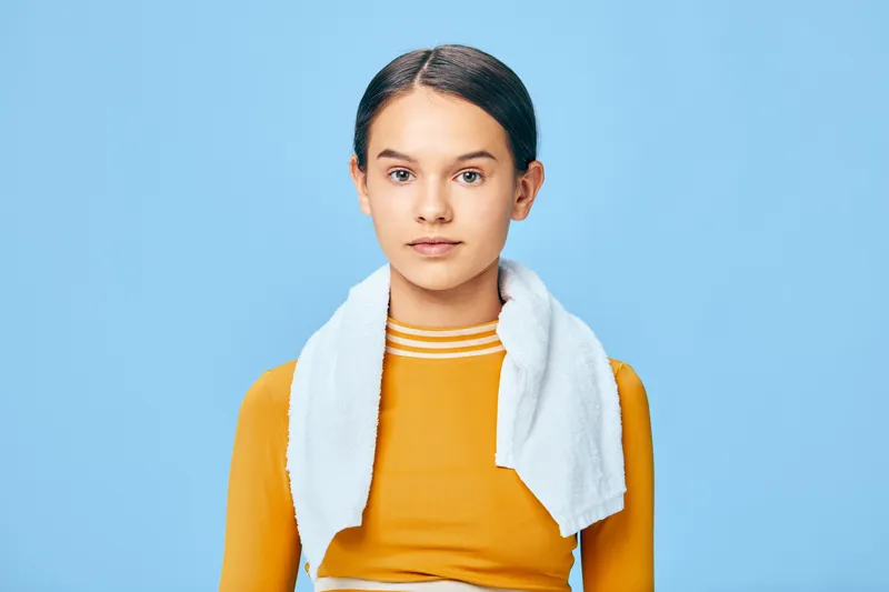 Woman fitness and sport concept with athletic pose, orange top and white towel around neck, blue studio background, emphasis on training, endurance and determination.