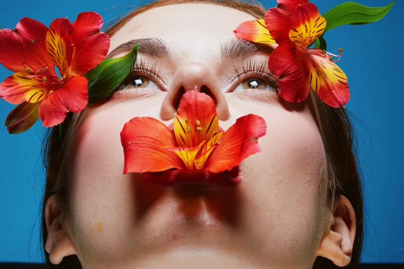 Young woman with vibrant flowers on her face, showcasing natural beauty and tranquility, against a bright blue background Emphasizes floral aesthetics and serenity