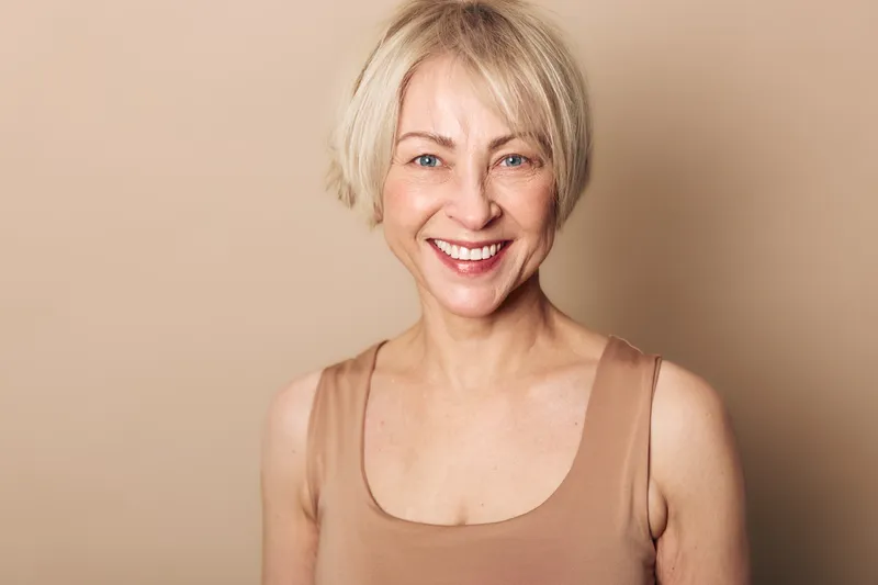 Smiling middle aged woman with short blonde hair wearing beige tank top stands against neutral background. Concept for happiness, confidence, natural beauty, and positive aging.