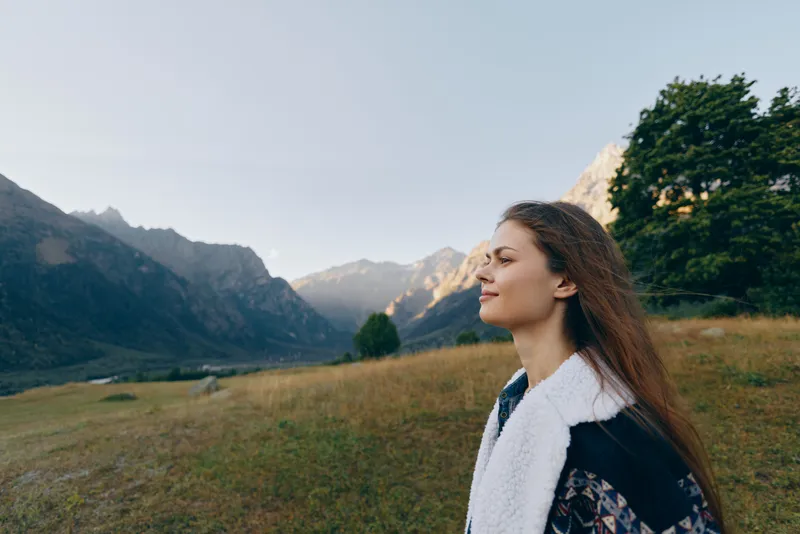 Woman profile portrait in mountains meadow, outdoors and contemplative with trees and distant peaks. Nature scene with calm expression, layered jacket and soft light over grassland.