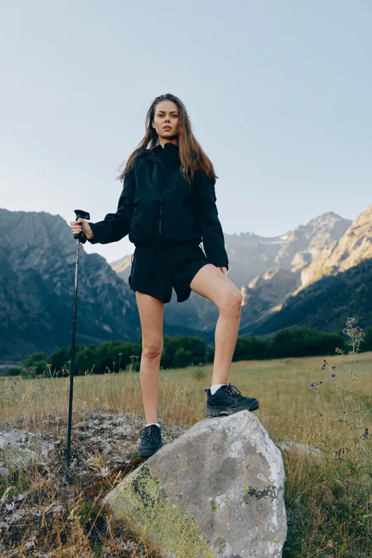 A confident woman stands on a rugged rock with a trekking pole in a wide mountain meadow under clear blue skies, embodying adventure, resilience, and exploration spirit.