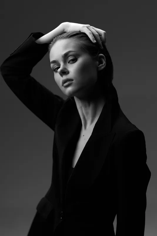 Black and white portrait of a young woman with hand on head wearing a dark suit, expressing calm and confidence in a studio setting