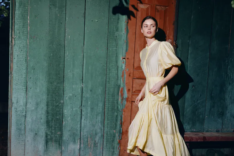 A fashionable woman in a yellow dress walks beside a rustic doorway in a vintage outdoor setting, exuding grace, elegance and timeless charm.