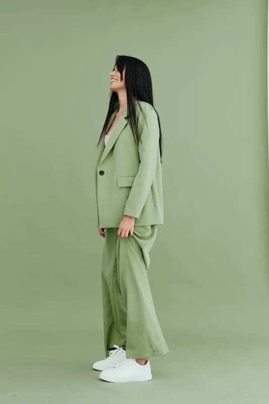 woman wearing green suit and white sneakers stands against green background with relaxed posture, showcasing modern casual fashion style and monochromatic palette.