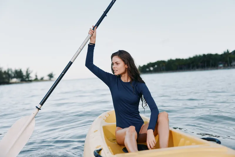 Young woman kayaking on calm water wearing a long sleeve shirt, enjoying outdoor adventure under clear sky with green forest in background.