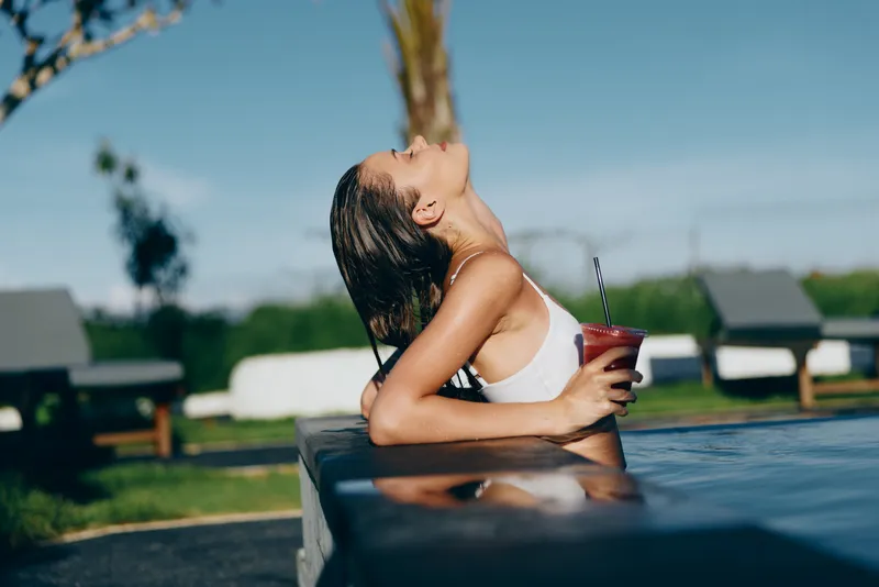 Outdoor pool scene with a woman enjoying relaxation, a refreshing drink, and a casual lifestyle moment under sunshine, breeze, and warm colors, portraying leisure and natural vitality