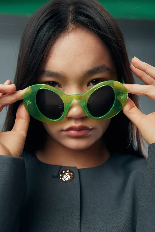 Young woman holding green sunglasses close to her face with serious expression. Fashion portrait with dark hair and plain background for style and eyewear concept.