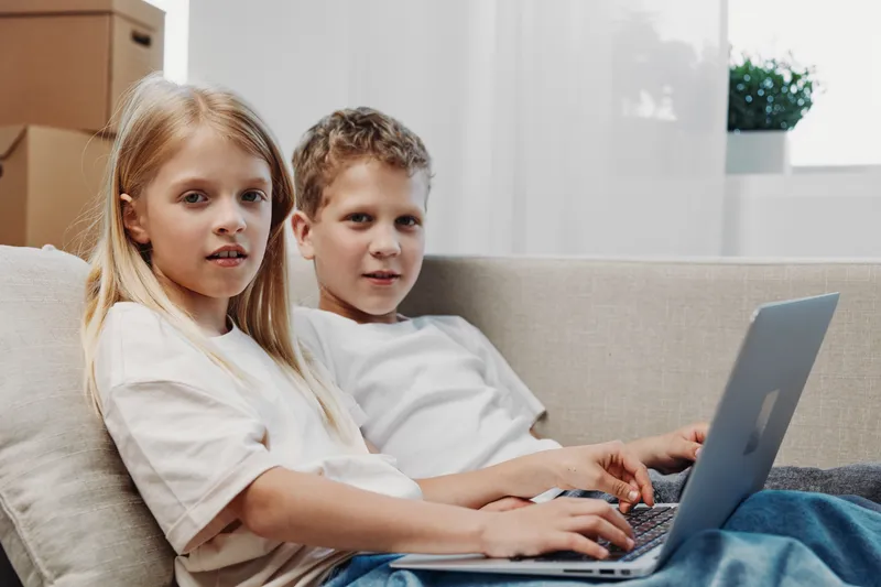 Two children sitting on a sofa using a laptop together, focused on screen in a bright modern living room environment.