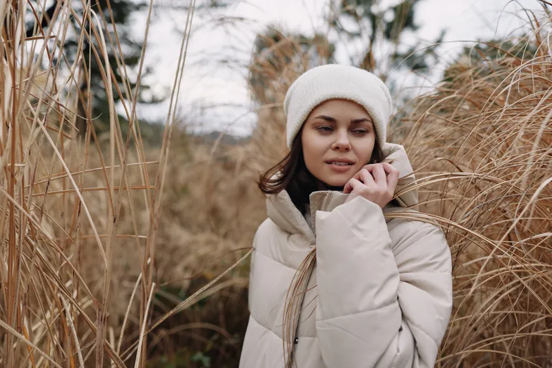 Woman in a warm coat talking on a phone amid tall dry grasses on a chilly day, outdoor portrait capturing calm mood and winter ambience in a natural setting