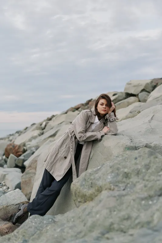 Young woman in long coat leans on large rocks under cloudy sky. Casual fashion outdoors with natural rocky background. Concept of autumn style and relaxed mood in nature.