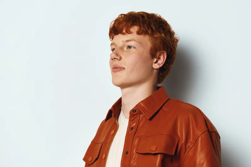 Young man with red hair wearing brown leather jacket and white shirt standing against white wall looking sideways with calm and confident facial expression in studio light.