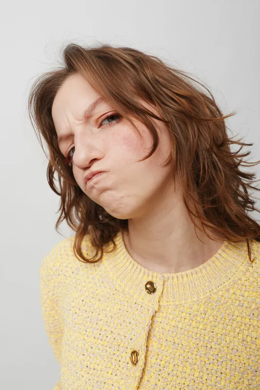 Young woman with messy brown hair wearing yellow sweater makes a displeased face with pursed lips and furrowed brows. Isolated on white background. Concept of frustration and annoyance.