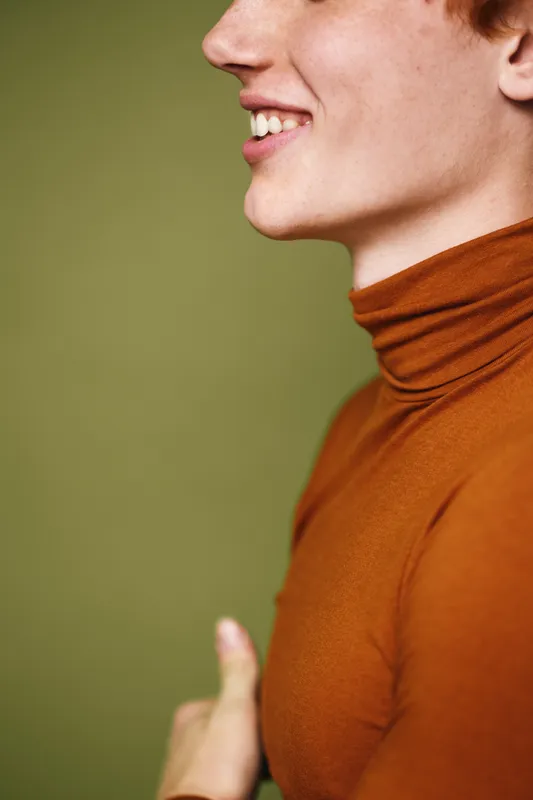 Profile of smiling young man wearing brown turtleneck sweater with thumbs up gesture on green background. Positive expression and casual clothing in closeup portrait.