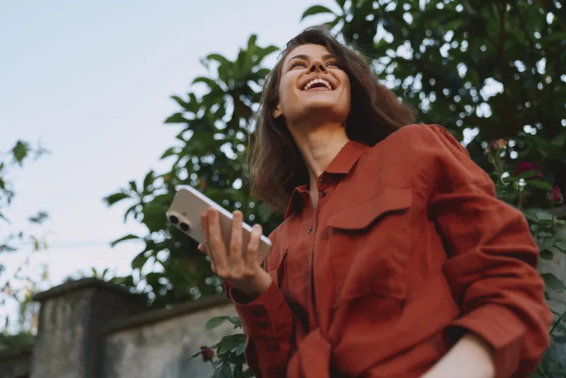 A smiling woman in a rust jacket enjoys the outdoors, holding a phone in one hand, with greenery and blue sky behind. Concept for lifestyle, optimism, and connection