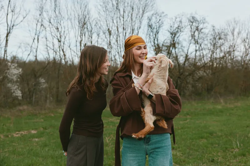 Two women outdoors with a smiling goat kid, sharing a playful moment in a grassy field. Lifestyle scene centered on friendship, care, and gentle animal bonding under soft natural light.