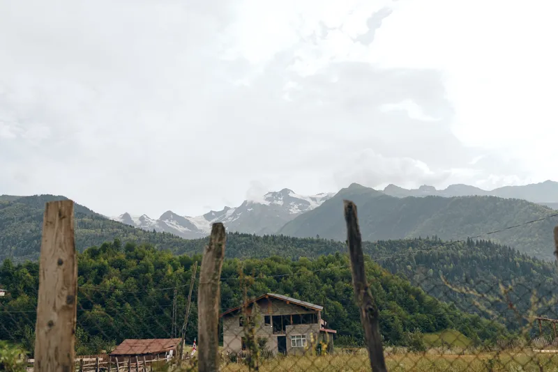 Landscape featuring distant mountains, a rustic cabin, and a weathered fence in the foreground, with green trees and a cloudy sky creating a tranquil countryside scene
