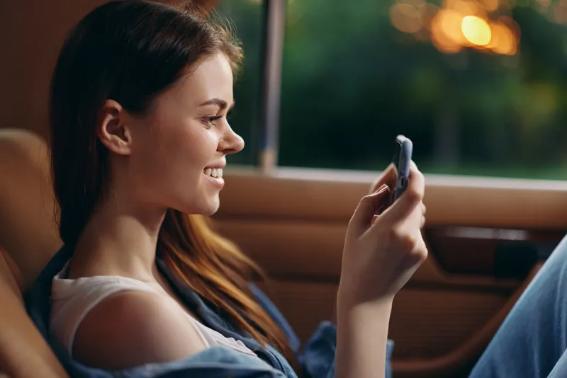 A young woman smiling while using a smartphone in a car, showcasing joy, casual attire, and a warm evening background with green hues and soft bokeh effects.