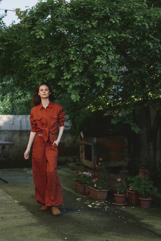 Candid woman lifestyle outdoors in a red orange dress, relaxed pose in a sunlit garden with lush greenery, path and pots, natural colors and tranquil mood