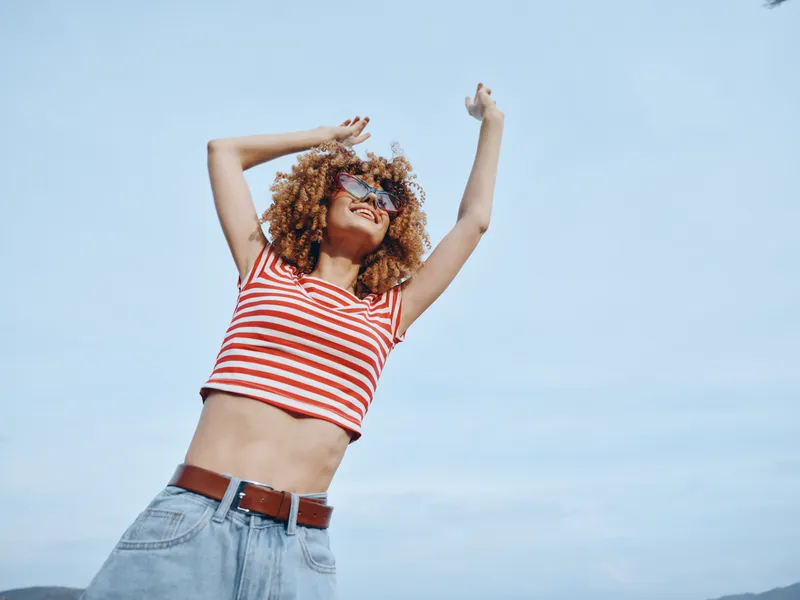 Joyful woman with curly hair dances outdoors under a bright blue sky, wearing a red striped crop top and denim jeans, expressing carefree summer energy.