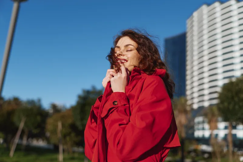 Confident young woman with brunette hair wears a bright red coat outdoors in an urban park, smiling softly as warm sunlight highlights modern buildings behind.