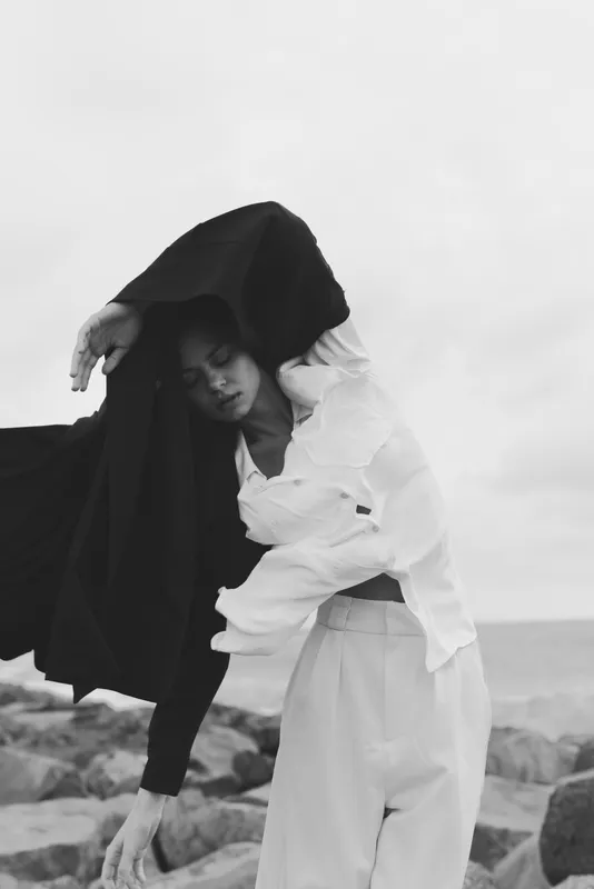 Cloak draped over a figure by the shore, dramatic pose, monochrome scene with ocean wind and rippled fabric. Fashion expression, mystery, and movement capture a balance between light, shadow, and