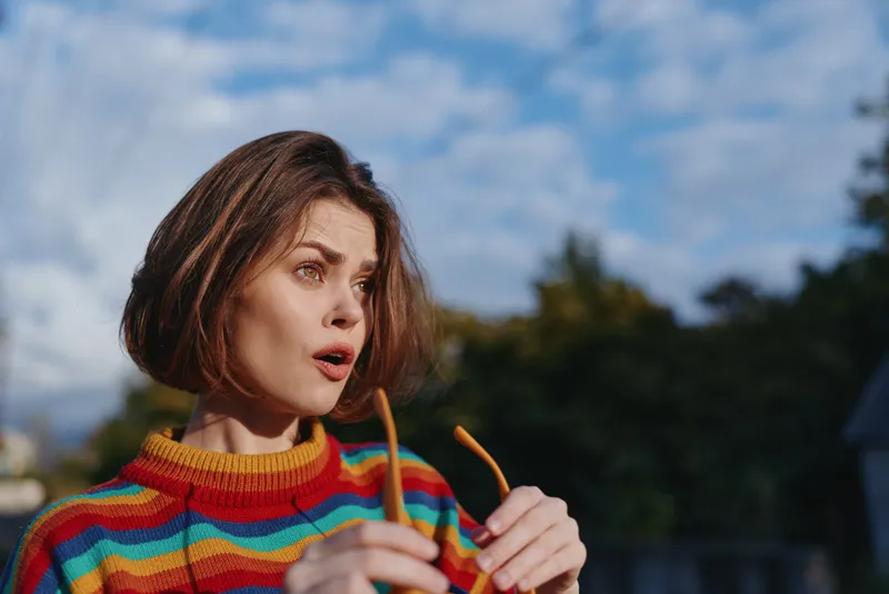 Woman surprised portrait of a young adult with short brown hair wearing a colorful striped sweater holding glasses outdoors, casual outfit and expressive travel lifestyle moment.