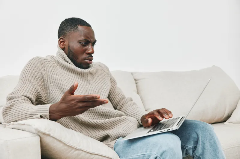 Young African man showing frustration while working on a laptop in a cozy, minimalist living room with neutral tones