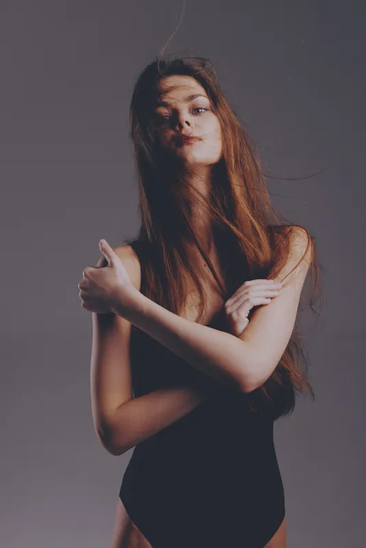Young woman with long hair posing confidently with crossed arms in moody lighting and minimalist background