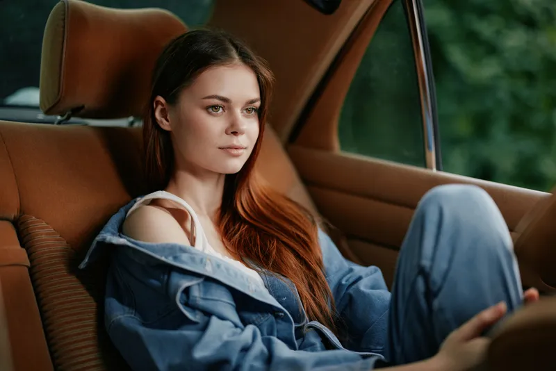Young woman with long red hair sitting in a car, wearing denim clothes, reflecting tranquility and comfort in a green natural background.