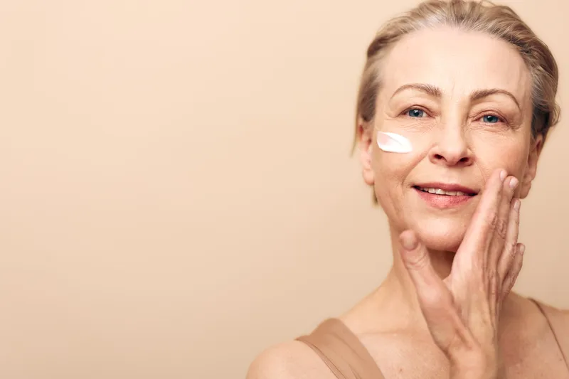 Mature woman with light skin applies moisturizing cream on cheek, smiling gently against beige background. Concept of skincare, aging care, beauty routine, and healthy skin hydration.
