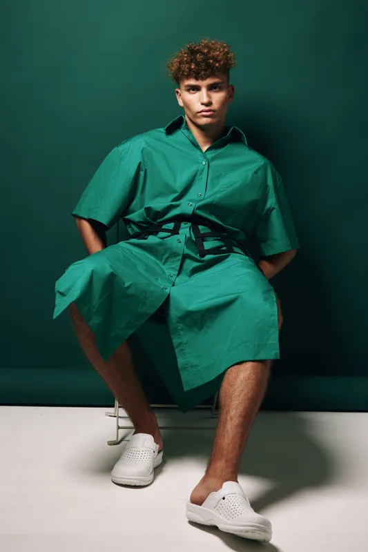 Young man with curly hair wearing green oversized shirt and shorts sitting on stool against dark green background. Casual fashion, relaxed pose and modern style with white slip-on shoes.