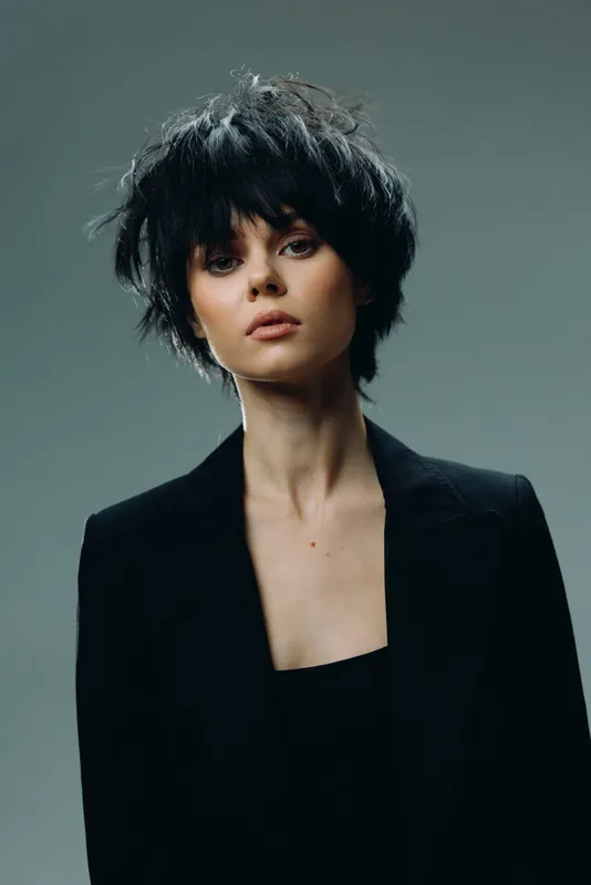 Stylish woman with short black hair wearing a dark blazer, looking confidently with neutral expression, minimalistic background