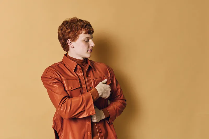 Young man with red hair wearing a brown leather jacket and plaid shirt posing with crossed arms on beige background in studio for fashion and style concept.