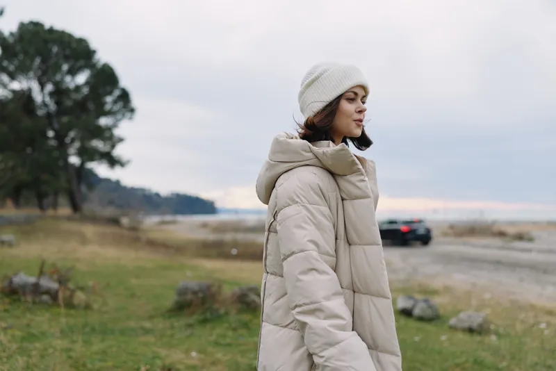 Woman in a beige puffer coat and knit beanie stands outdoors by a coastal field under an overcast sky, with distant water and a blurred road
