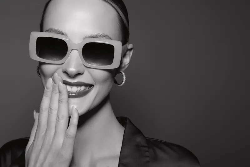 Smiling young white woman with pale skin wearing large square sunglasses and hoop earrings covers mouth with hand. Black and white studio portrait with elegant mood and fashion style.
