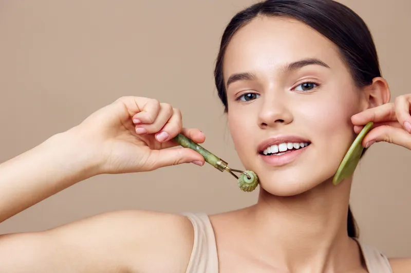 Young woman using jade roller for skincare on neutral background, promoting healthy skin and beauty routines Emphasizes wellness and natural beauty