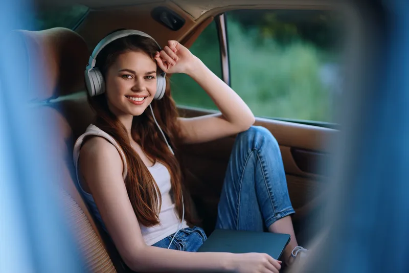 Young woman enjoying music in a car, wearing casual clothing and headphones, smiling happily, with a natural outdoor background emphasizing relaxation and joy.