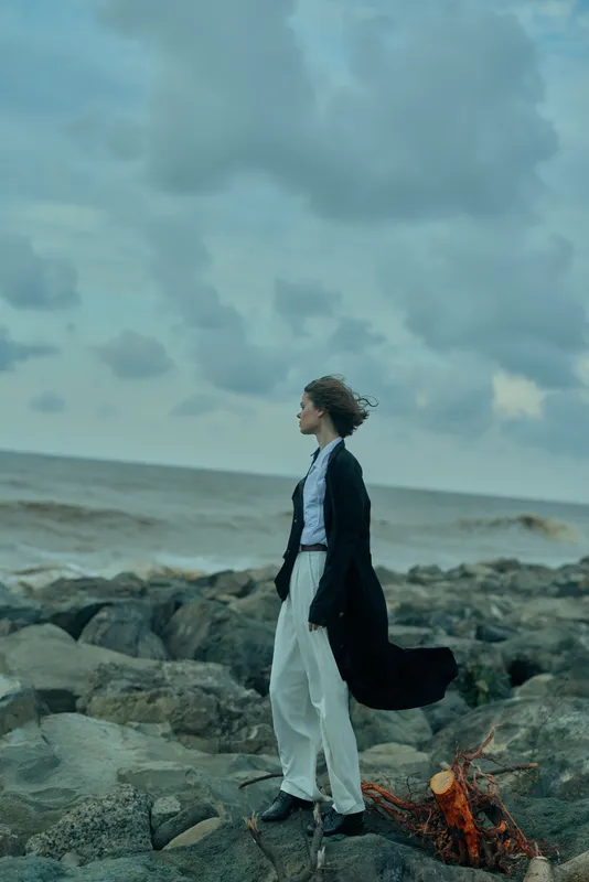 Wind swept woman in smart white pants and dark coat on rocky shore facing the sea, blue sky, dramatic mood and outdoor fashion by the ocean